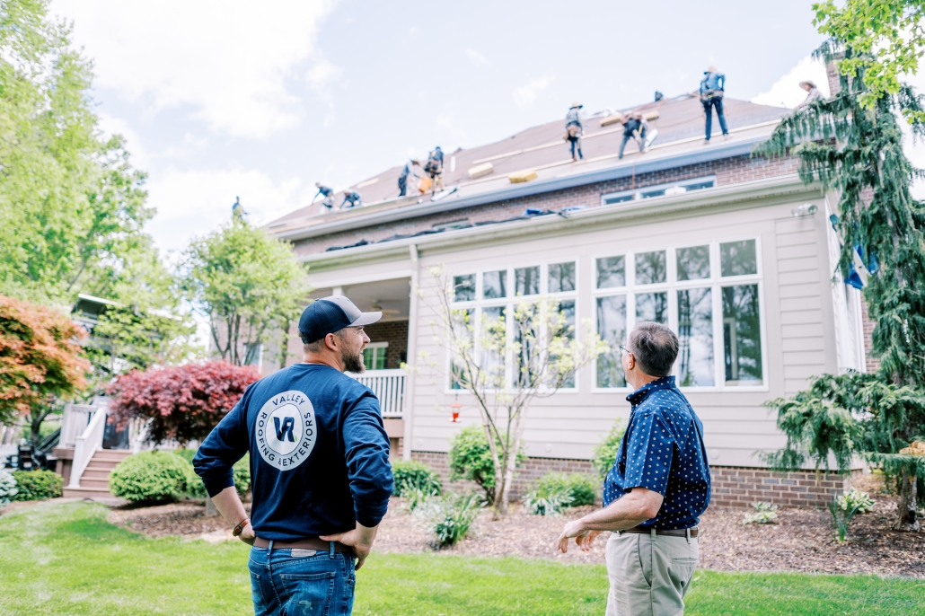 professional photo of roofing company employee interacting with a customer in lawn while roofers install new roof on home.